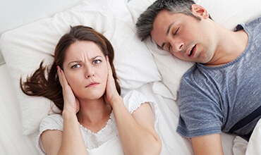 Woman covering her ears in bed next to snoring man
